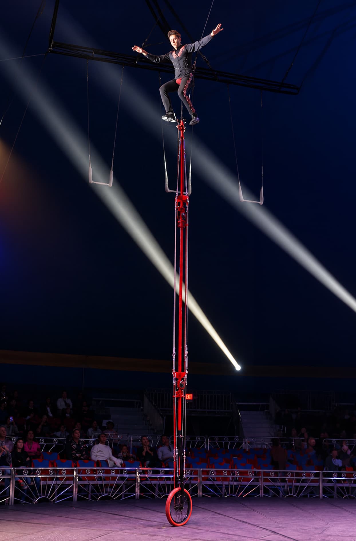A performer balancing on top of a 27-foot unicycle, arms out, far above the arena floor