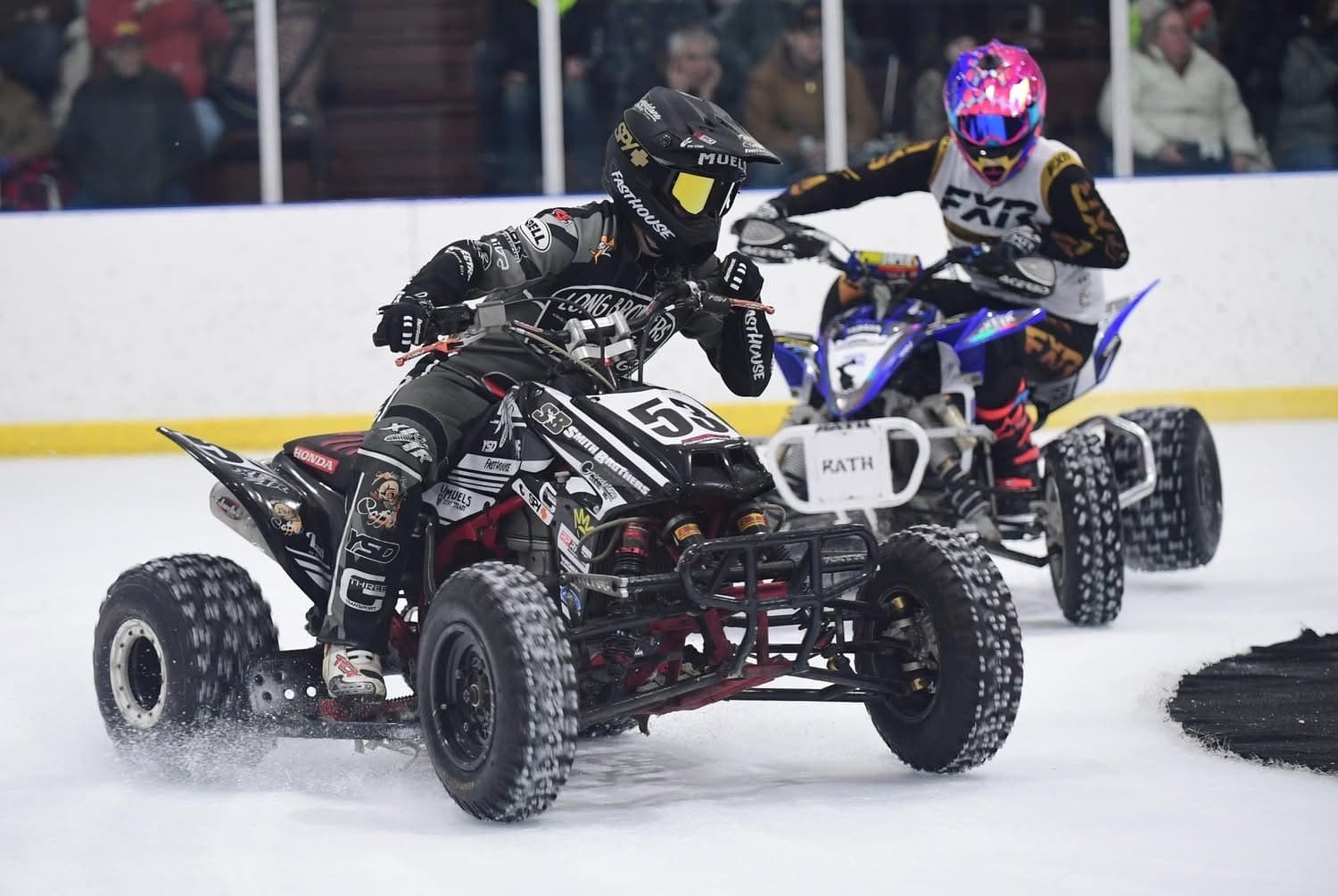 Two racing quads side by side on arena ice, throwing ice from their studded tires